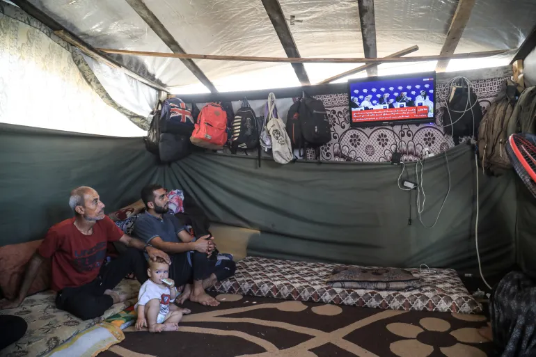 Wael al-Nabahin dan keluarganya menonton televisi dalam tenda di lokasi pengungsian Deir el-Balah, kawasan Gaza Tengah. @Al Jazeera